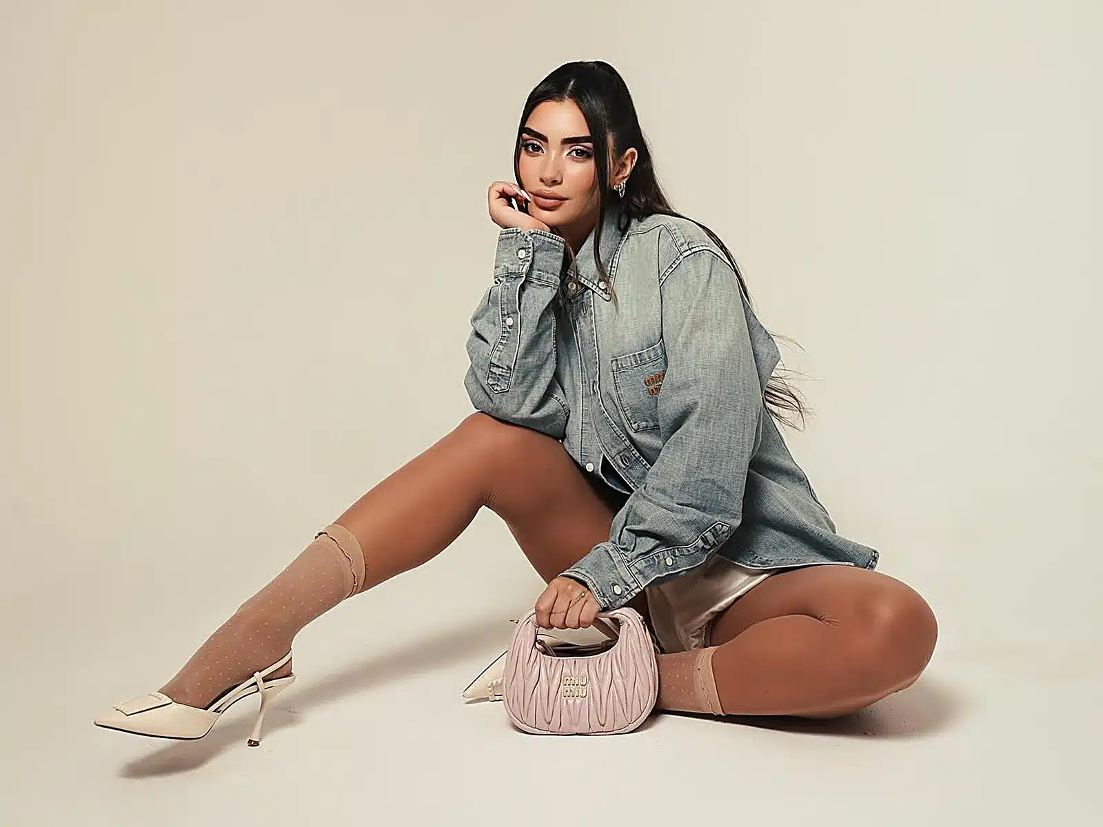 Woman in an oversized denim shirt seated on the studio floor beside a pink designer handbag at a Miami photo studio