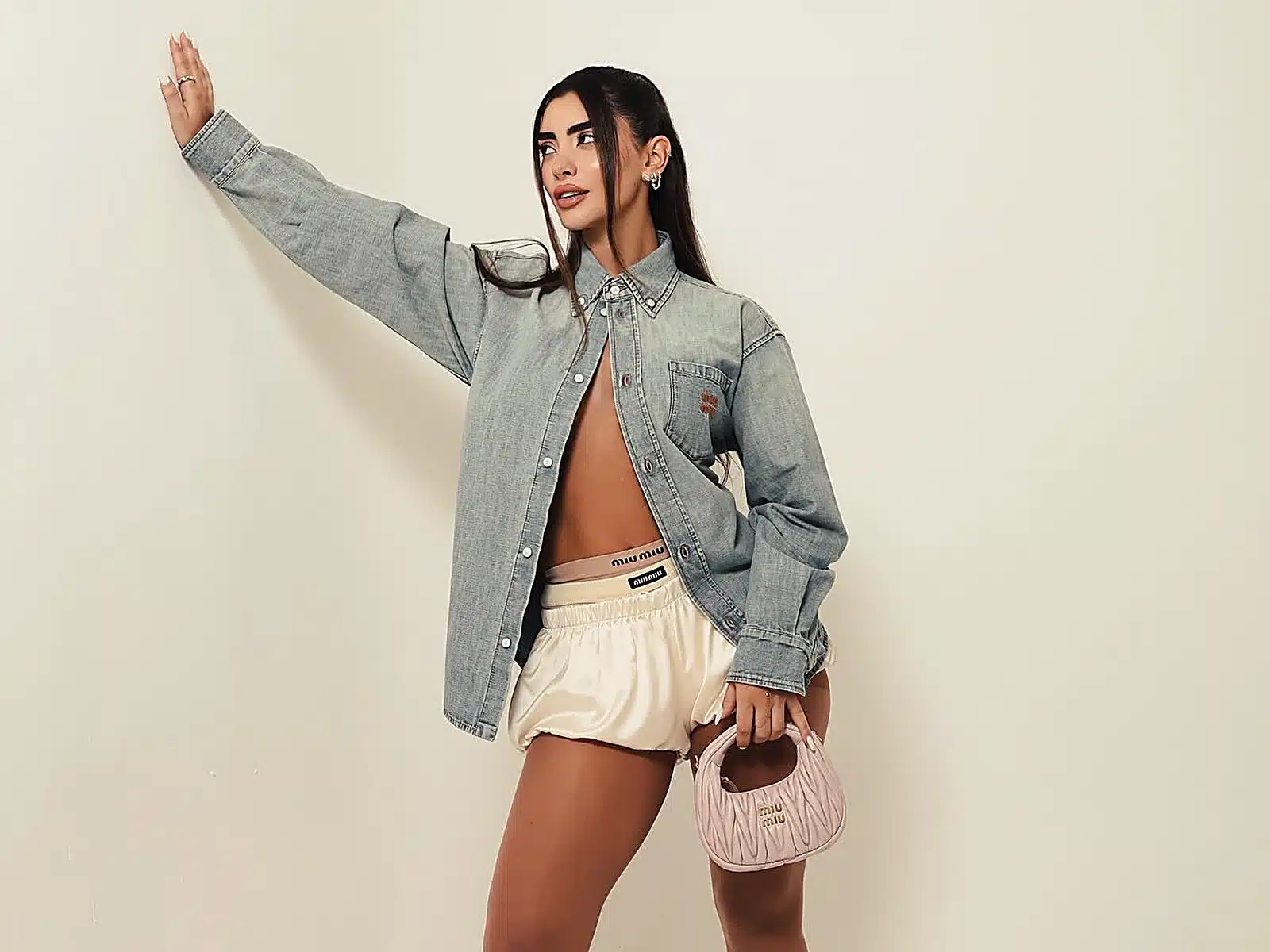 Woman in an oversized denim shirt and satin shorts leaning against a wall holding a pink designer handbag at a Miami photo studio