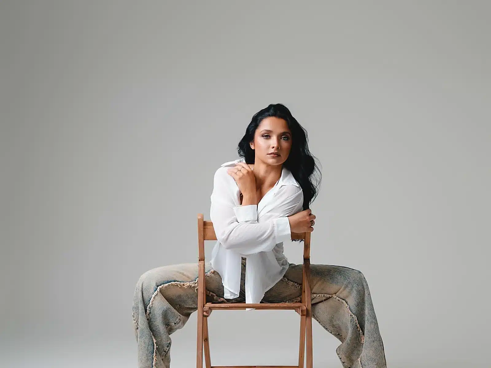 Woman in a white shirt and denim jeans posing seated on a wooden folding chair in a minimalist studio at a Miami photo studio