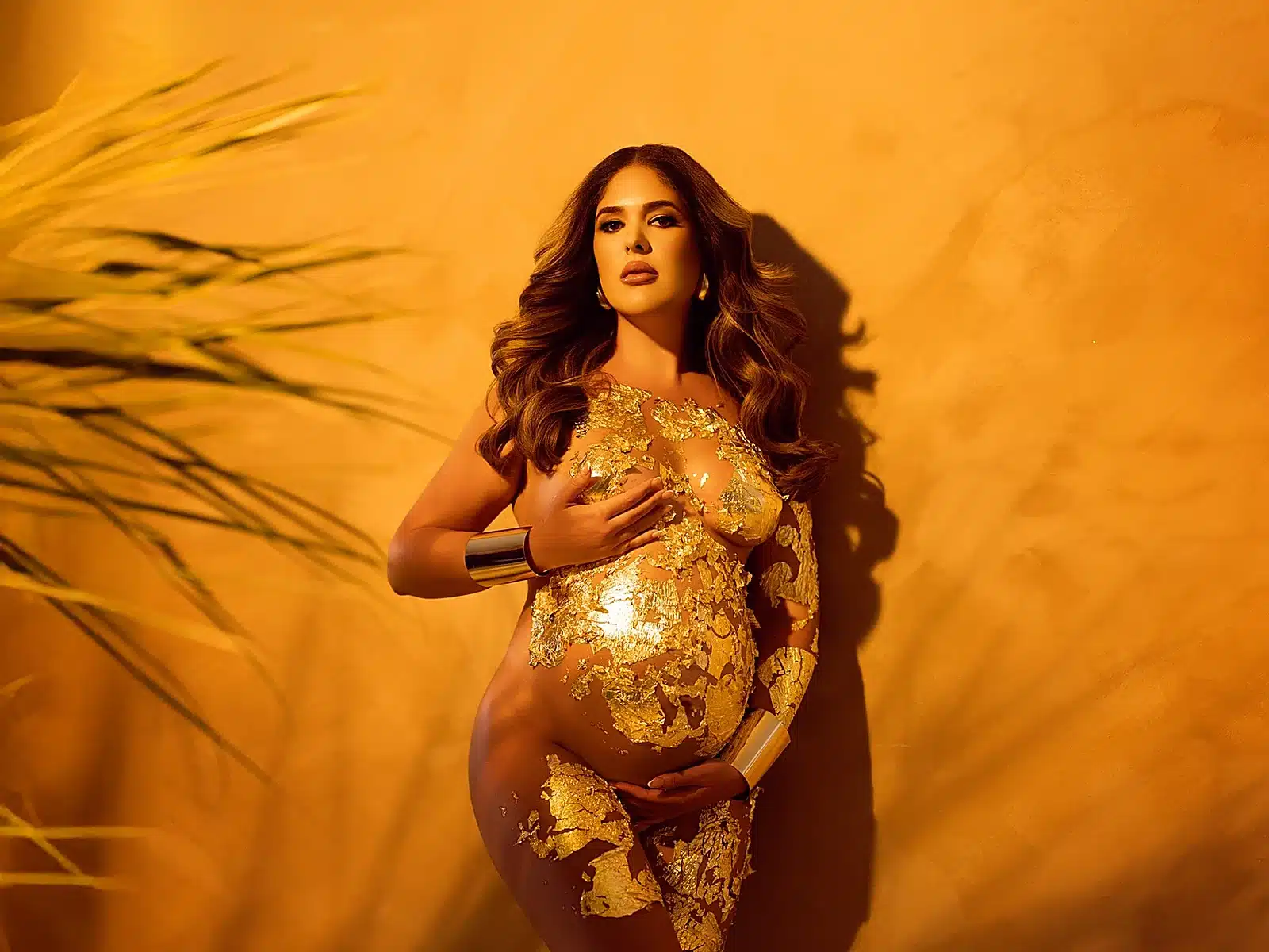 Pregnant woman in a gold-leaf bodysuit posing against a warm amber wall in an artistic maternity shoot at a Miami photo studio