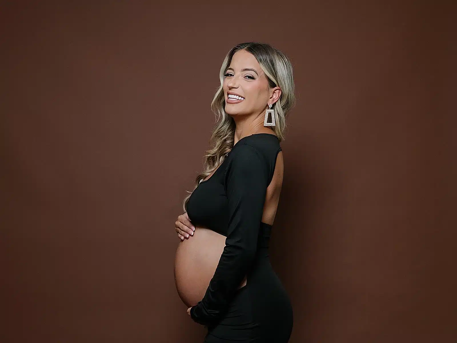 Smiling pregnant woman in a black gown holding her belly against a brown studio backdrop at a Miami photo studio